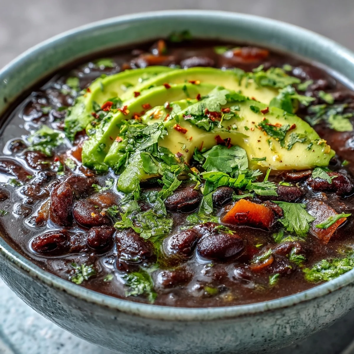 A comforting bowl of Black Bean Soup garnished with creamy avocado and fresh cilantro, steaming beside a lime wedge on a rustic wooden table.