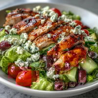 A colorful Greek salad bowl with juicy grilled chicken, fresh veggies, and creamy feta on a white plate.